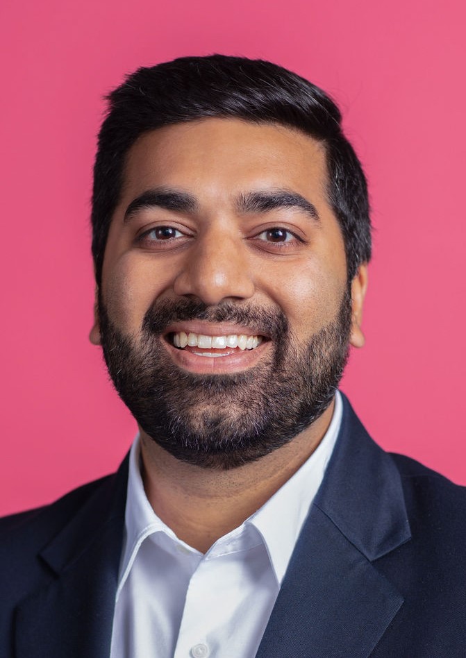 An indian man smiling in front of a pink background.